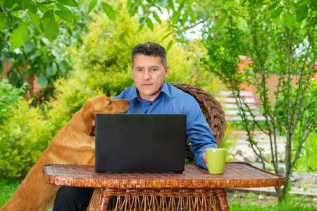 Mature Caucasian male with his dog is drinking coffee and working with a laptop in the home garden. Remote business conceptの写真素材