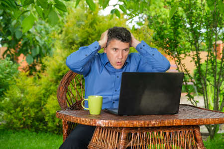 Shocked businessman reading newspaper with fake news while sitting near laptop in his backyard garden. Excessive stress at work. Stressful work environmentの写真素材