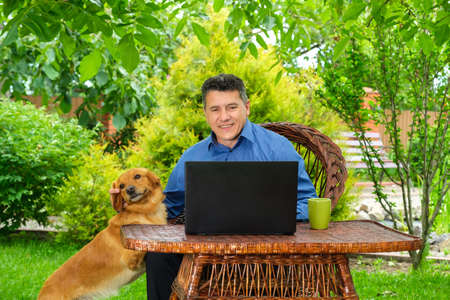 Mature Caucasian Man works and reading news via laptop and petting a dog in the backyard garden. Remote business conceptの写真素材