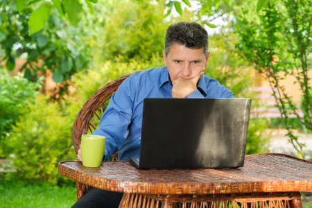 Serious middle-aged businessman drinking coffee and reading news using laptop sitting in his backyard garden. Remote working conceptの写真素材