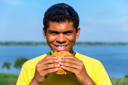 Hungry smiling African American man enjoying the taste of hamburger outdoor in summer. Concept of...