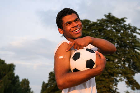Young smiling Afro American sportsman holds soccer ball and showing his arm after receiving a vaccine at sunset in summerの写真素材