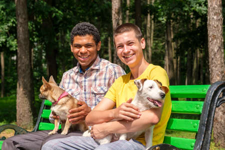 Two smiling african american and caucasian friends with dogs relaxed on the bench in park in summerの写真素材