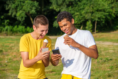 Two young cheerful and laughing African-American and caucasian friends eats ice cream and use smartphone, makes selfie while standing in park outdoors in summerの写真素材