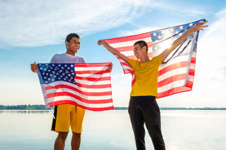 Two young cheerful and laughing African American and Caucasian friends proudly holding an American flag against the blue sky at sunset in the summerの写真素材
