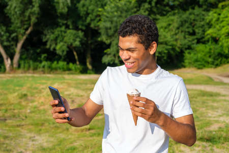 Young cheerful and laughing African-American and Caucasian man eats ice cream and use smartphone, makes selfie while standing in park outdoors in summerの写真素材
