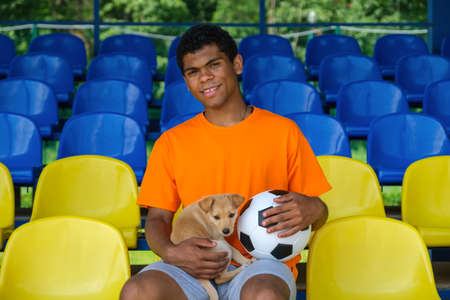 Young African American guy with a soccer ball and dog sitting on an empty football grandstand outdoor in summerの写真素材