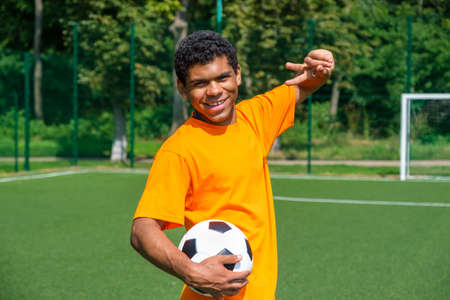 Portrait of young smiling African-American man holding soccer ball and showing victory sign while standing on sports court outdoors against soccer goalの写真素材
