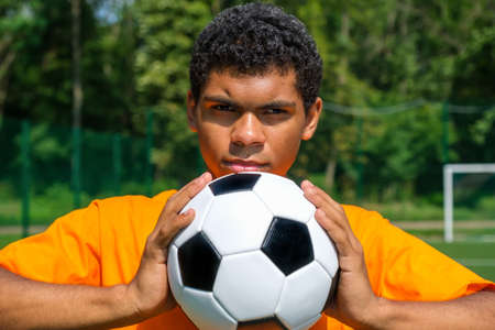 Portrait of young smiling African-American man holding soccer ball close up while standing on sports court outdoors against soccer goalの写真素材