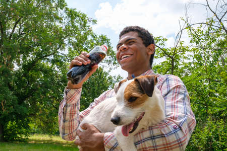 Young smiling cheerful African American man holding Jack Russell terrier dog and drinking cola from bottle outdoors. Walking in park in summer, low angle viewの写真素材