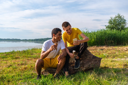 Two young cheerful and laughing African-American and caucasian friends eats ice cream and use smartphone, makes selfie while sitting on beach of river outdoors in summerの写真素材