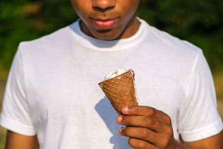 Ice cream cone in hand of young African American man close up. Guy eats melted ice cream outdoors in summer.の写真素材