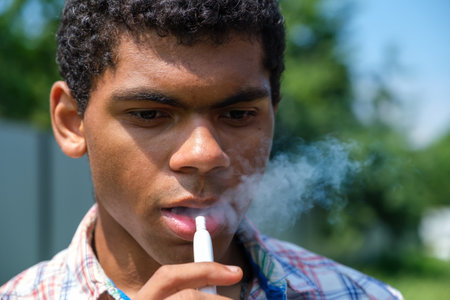 Close-up Brazilian young man vaping e-cigarette with e-liquid and breathes out steam or vapor outdoors in summer. Vaping conceptの写真素材