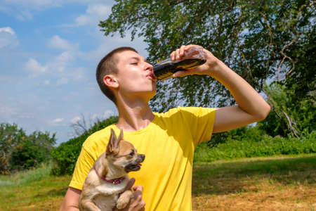 Young caucasian man holding Chihuahua dog and drinking cola from bottle outdoors. Walking in park in summer, low angle viewの写真素材