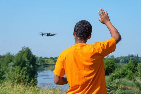 Young smiling African American man operating drone with controller standing with back and waving with his hand on the meadow near the river.の写真素材
