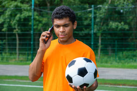 Portrait of young Brazilian man holding soccer ball and use smartphone while standing on sports court outdoors against soccer goalの写真素材