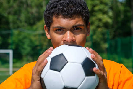Portrait of young Brazilian man holding soccer ball close up while standing on sports court outdoors against soccer goalの写真素材