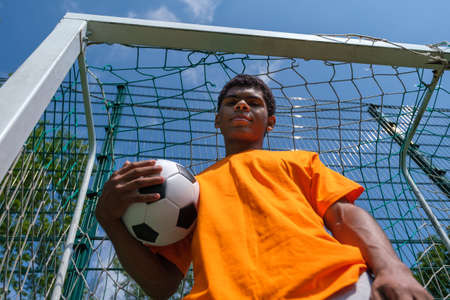 Young Brazilian man holding soccer ball while standing in soccer goal on sports court outdoors in summer, bottom viewの写真素材