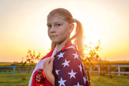 Portrait of a cute little girl, wrapped in an American flag, standing in a field at sunset in summer. American federal holidays, independence day conceptの写真素材