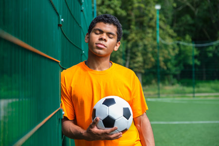 Side view portrait of young Brazilian man holding soccer ball while standing against fence in sports court outdoors, copy spaceの写真素材