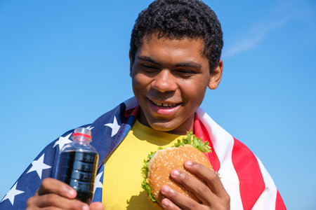 Hungry African American man enjoying the taste of hamburger and cola drink while sitting wrapped in american flag outdoor in summer close-up. Concept of love to junk food dietの写真素材