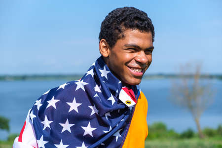 Young smiling African American man proudly holding American flag covered on his shoulders against blue sky in summerの写真素材