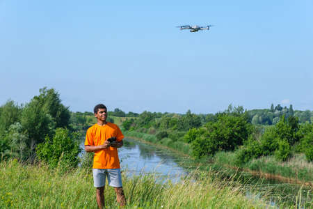 Full length portrait of young smiling African American man operating drone with controller in hands on the meadow near the river.の写真素材