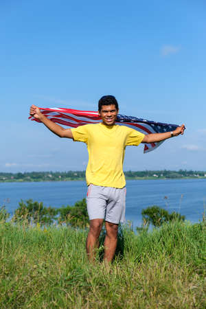 Full length young African American man standing with outstretched arms with USA flag by the river on blue sky background in sunny summer weather. Copy space for text, vertical orientationの写真素材