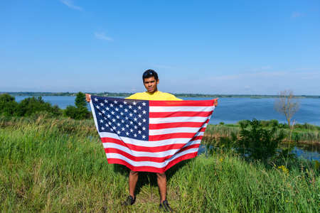 Full length young African American with USA flag standing by the river on blue sky background in sunny summer weather. Copy space for textの写真素材