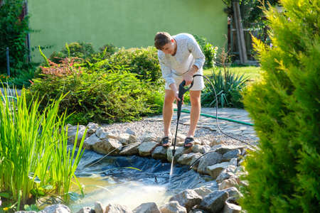 Young caucasian man cleans artificial garden pond bottom with high-pressure washer nozzle from mud and sludge. Spring and summer pond care workの写真素材