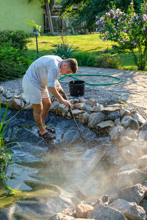 Young caucasian man cleans artificial garden pond bottom with high-pressure washer nozzle from mud and sludge. Spring and summer pond care work. Vertical orientationの写真素材