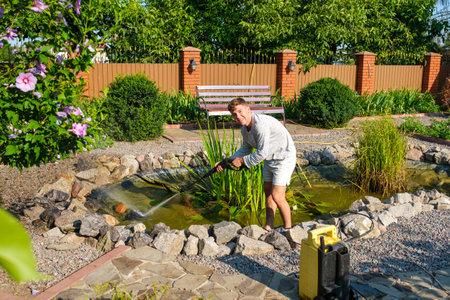 Young smiling caucasian man cleans artificial garden pond bottom with high-pressure washer nozzle from mud and sludge. Spring and summer pond care workの写真素材
