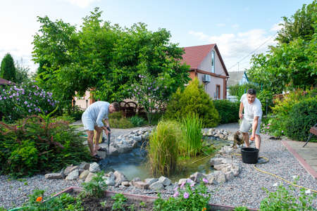 Mature father and young adult son cleaning artificial garden pond bottom with high-pressure washer nozzle from mud and sludge. Spring and summer pond care workの写真素材