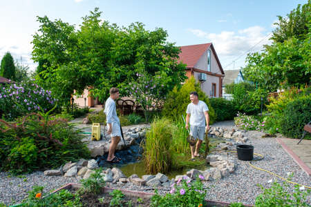 Mature father and young adult son cleaning artificial garden pond bottom with high-pressure washer nozzle from mud and sludge. Spring and summer pond care workの写真素材