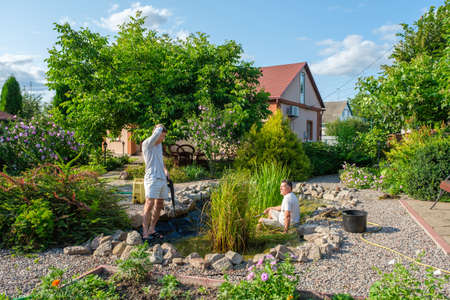 Mature father and young adult son cleaning artificial garden pond bottom with high-pressure washer nozzle from mud and sludge. Spring and summer pond care workの写真素材
