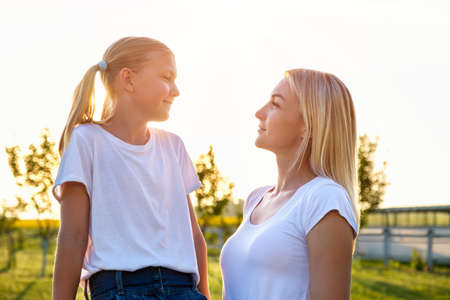 Portrait of caucasian mother and daughter looks on each other in countryside in sunset in summer. Parenthood, childhood, love, relations concept.の写真素材