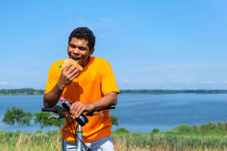 Hungry young African American man eating a hamburger and drinking cola from a bottle on a bicycle ride along the river on a background of blue sky in sunny summer weather. Copy space for textの写真素材