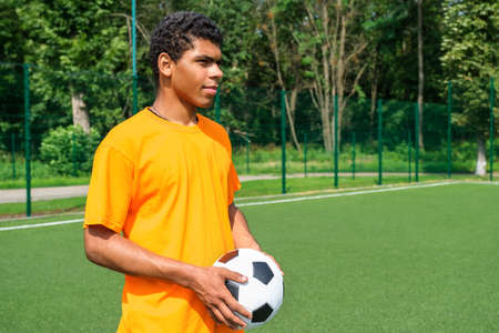 Portrait of young smiling Brazilian man holding soccer ball while standing on sports court outdoors against soccer goalの写真素材