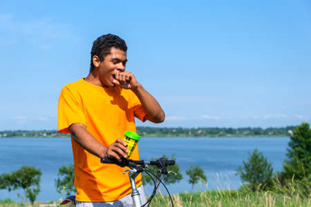 Young African American man drinking coffee and yawning on a bicycle ride along the river, on a background of blue sky, in sunny summer weather. Copy space for textの写真素材