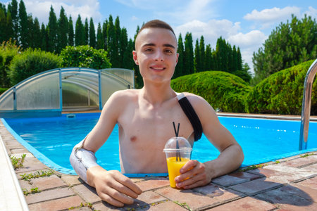 Young smiling guy with an amputated arm and wearing a prosthesis drinks a cocktail in swimming pool in the courtyard of his houseの写真素材
