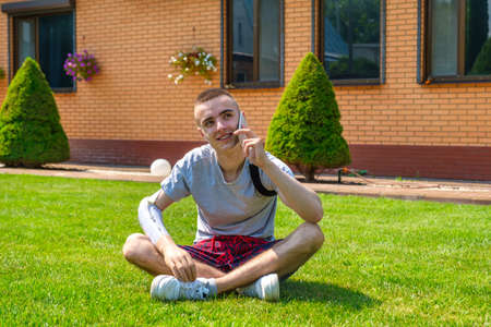 Young smiling caucasian guy with amputated arm and prosthesis talking on the phone while sitting on the front lawnの写真素材
