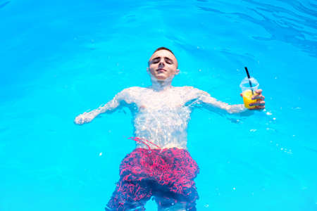 Young caucasian guy with an amputated arm floating in a swimming pool with blue water and holds in his hand a cocktailの写真素材