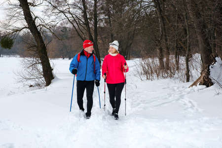Smiling senior couple walking with nordic walking poles in snowy winter forest. Elderly wife and husband doing healthy exercise outdoors. Active lifestyle after retirement concept.の写真素材