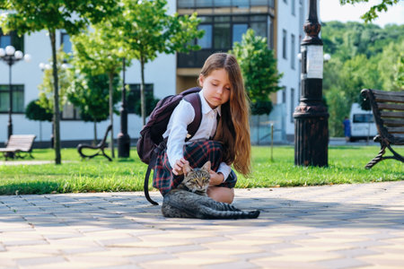 Cute little girl in school uniform and with backpack stroking a stray cat on the street, in the first time goes to school.の写真素材