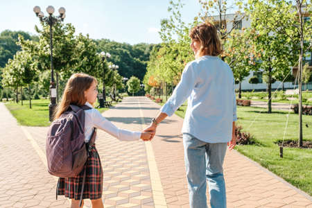 Daughter in school uniform and mother walking together to school down the street, holding hands, back viewの写真素材