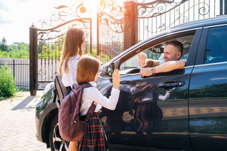 Smiling middle-aged father in the car, leaving her daughters and sisters in school uniform at school and gesturing goodbye.の写真素材