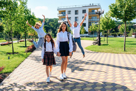 Two sad girls sisters in school uniforms hold hands and go to school together while their parents jump for joy in the morning, front viewの写真素材