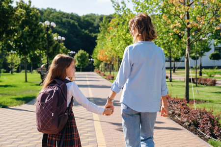Daughter in school uniform and mother walking together to school down the street, holding hands, back viewの写真素材