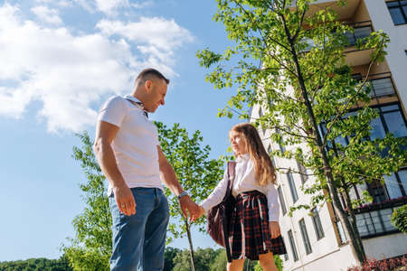 Back to school. Caucasian schoolgirl with a backpack and uniform holds her smiling father by the hand in the street before going to school.の写真素材