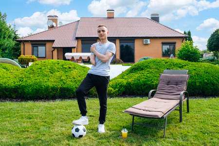 Full length young caucasian man with amputated arm, holds foot on soccer ball while standing on the lawn in the backyard of his home, disability rehabilitation and active lifestyleの写真素材